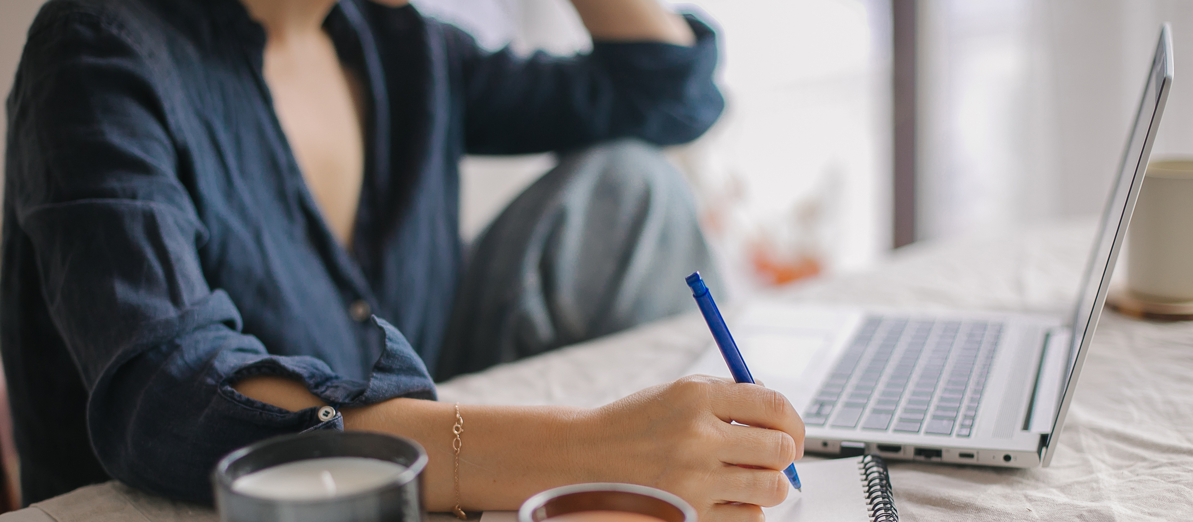A photo of woman sitting at a computer at her desk writing notes in a notebook